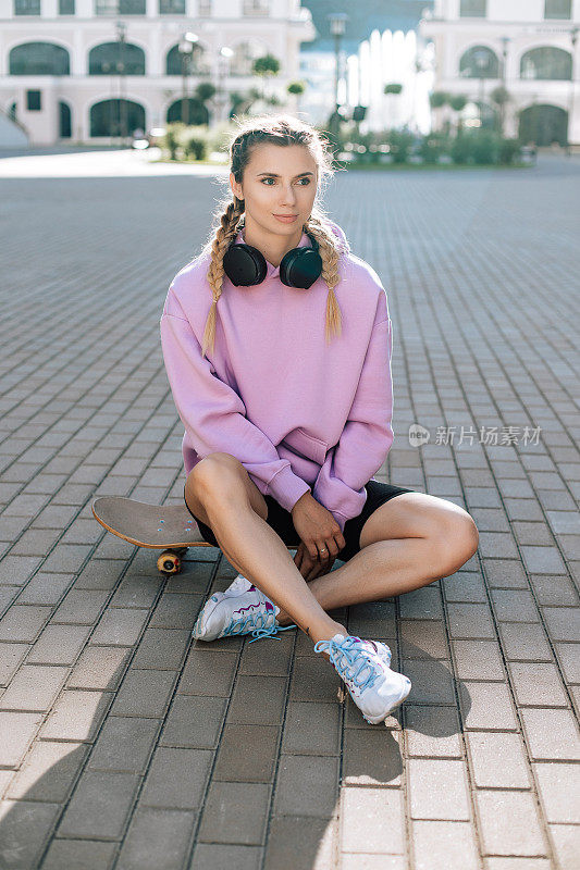 Portrait of woman with wireless black headphones who enjoying and listening music outdoors the street. Girl is sitting on skateboard.